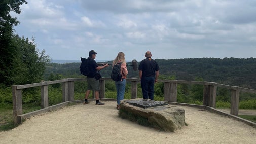 Three people with their backs to the camera enjoying the view at Hindhead Commons. The ranger is being interviewed by a woman in a pink jacket and a sound recordist is standing next to her.